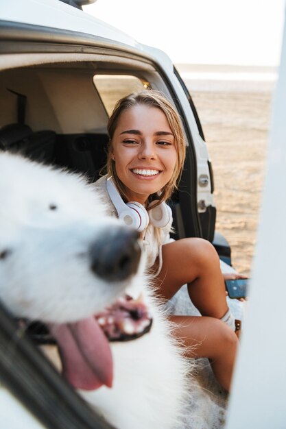 Imagen de mujer joven positiva abrazando perro samoyedo al aire libre en la playa en coche.