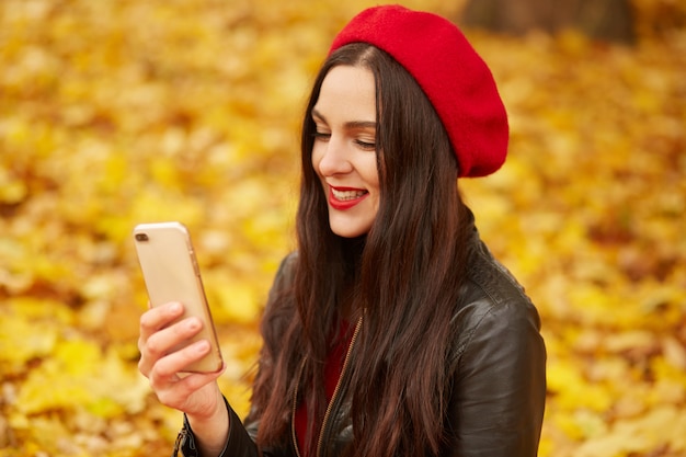 Imagen de la mujer joven que hace el selfie o que tiene videollamada en parque del otoño. Hermosa chica viste redberet