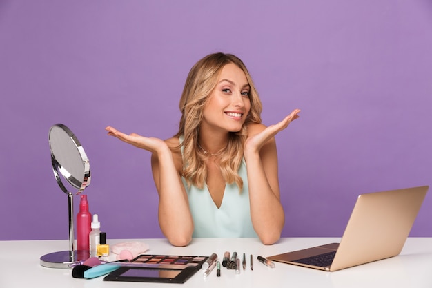 Imagen de mujer joven sonriente usando laptop mientras hace maquillaje y sentado en el escritorio aislado