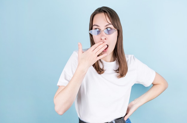 Imagen de mujer joven sorprendida aislada sobre fondo azul con anteojos azules, cubre su boca con la mano derecha