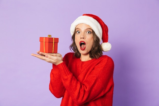 Imagen de una mujer joven sorprendida emocionada que lleva el sombrero de la Navidad aislada sobre la pared púrpura que sostiene la caja de regalo sorpresa.