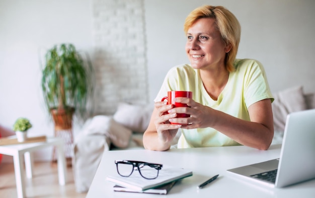 Foto imagen de una mujer madura feliz y tranquila con ropa casual con una bebida en las manos mirando hacia otro lado mientras está sentada en un escritorio en la cocina de casa