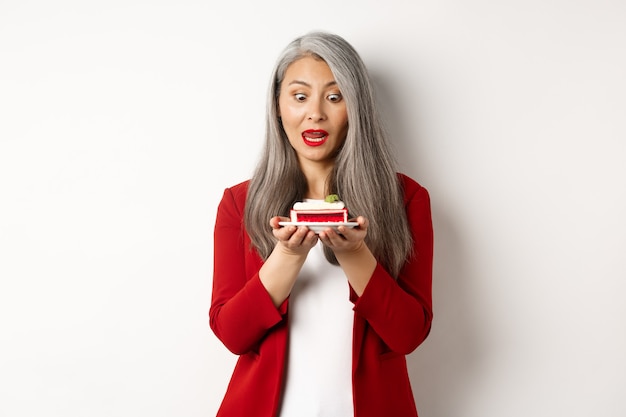 Imagen de una mujer mayor asiática divertida que mira tentada al pedazo de la torta, el deseo de morder el postre, de pie sobre el fondo blanco.