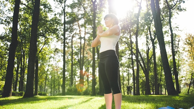 Imagen de mujer de mediana edad sonriente en ropa de fitness haciendo ejercicios de estiramiento y yoga. Mujer meditando y haciendo deportes en la estera de fitness sobre el césped en el parque