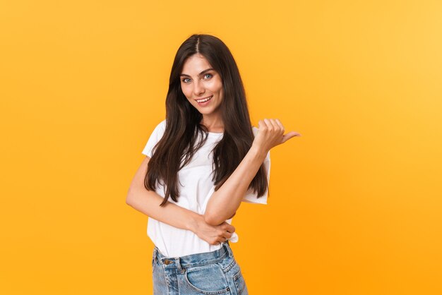 Imagen de mujer morena caucásica con cabello largo sonriendo y apuntando con el dedo en copyspace aislado sobre pared amarilla