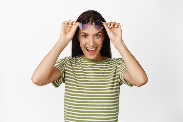 Imagen de una mujer morena feliz quitándose las gafas de sol y mirando emocionada a la cámara mirando algo interesante de pie con ropa de verano sobre fondo blanco