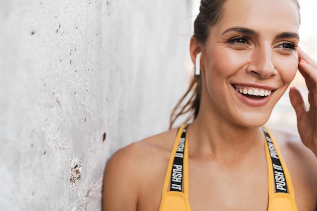 Foto imagen de mujer morena sonriendo y tocando earpods mientras está parado sobre un muro de hormigón al aire libre