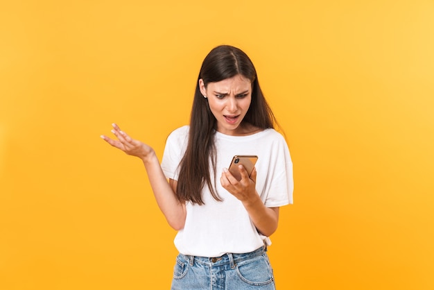 Imagen de mujer morena tensa con ropa básica que expresa descontento mientras sostiene el teléfono celular aislado sobre la pared amarilla