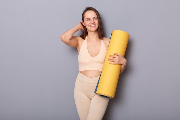 Imagen de una mujer muy linda y encantadora con top beige y leggins sosteniendo una alfombra de fitness aislada sobre un fondo gris tocando su cabello sonriendo a la cámara visitando el gimnasio para mejorar la forma del cuerpo