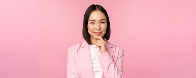 Imagen de una mujer de negocios asiática parada en una pose de pensamiento haciendo una lluvia de ideas con traje Empresaria vendedora coreana posando sobre un fondo rosa