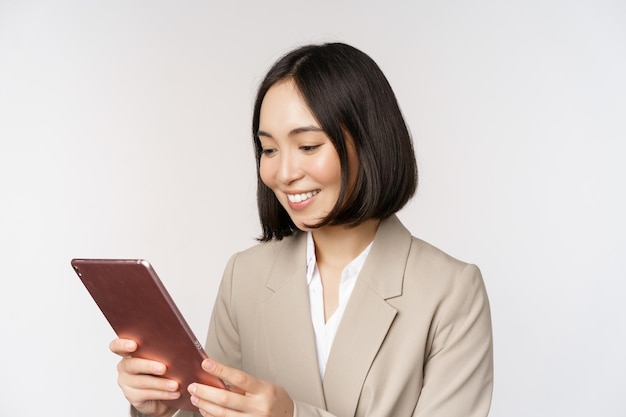 Imagen de una mujer de negocios asiática usando una tableta digital mirando un aparato y sonriendo trabajando de pie contra un fondo blanco