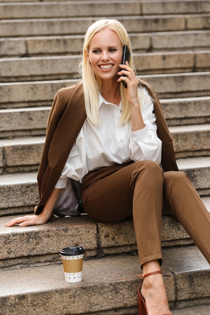 Imagen de una mujer de negocios asombrosa joven alegre feliz sentarse en pasos al aire libre en ropa formal con café hablando por teléfono móvil.