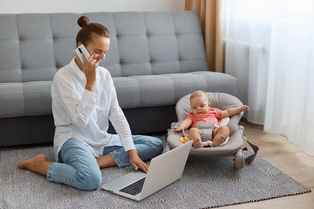 Imagen de una mujer con peinado de moño con camisa blanca y jeans sentada en el suelo cerca de la tos con un bebé en una mecedora mujer conversando por teléfono móvil y escribiendo en un cuaderno