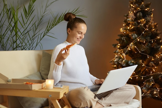 Imagen de una mujer positiva y adorable trabajando en una laptop en casa en Navidad sentada tosiendo cerca del árbol de Navidad con luces trabajando en línea durante las vacaciones de invierno y comiendo pizza
