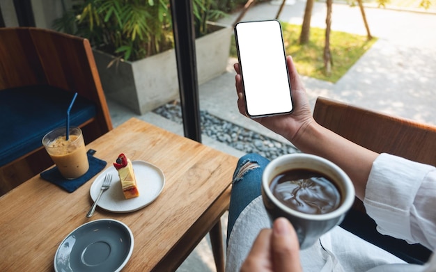 Imagen de una mujer que sostiene un teléfono móvil con una pantalla de escritorio blanca en blanco mientras bebe café en un café