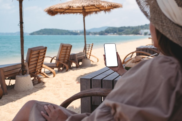 Imagen de una mujer que sostiene un teléfono móvil con una pantalla de escritorio en blanco mientras está sentada en la playa