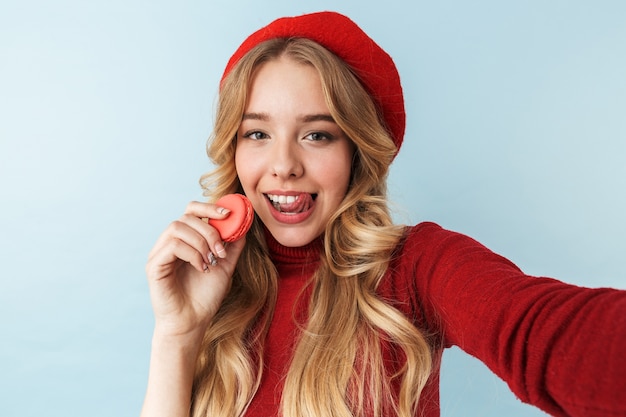 Imagen de mujer rubia europea de 20 años vistiendo boina roja comiendo galletas de macarrón mientras toma una foto selfie, aislado