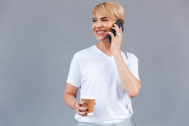 Imagen de mujer rubia satisfecha con camiseta blanca de pie aislado sobre una pared gris y sosteniendo café de una taza de papel mientras habla por teléfono celular