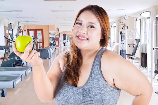 Imagen de una mujer con sobrepeso sonriendo a la cámara mientras sostiene una manzana en el gimnasio