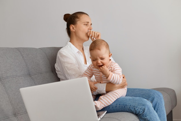 Imagen de mujer soñolienta con peinado moño vestida con camisa blanca y jeans sentada en el sofá con su bebé y bostezando, trabajando largas horas en la computadora portátil y cuidando al niño.