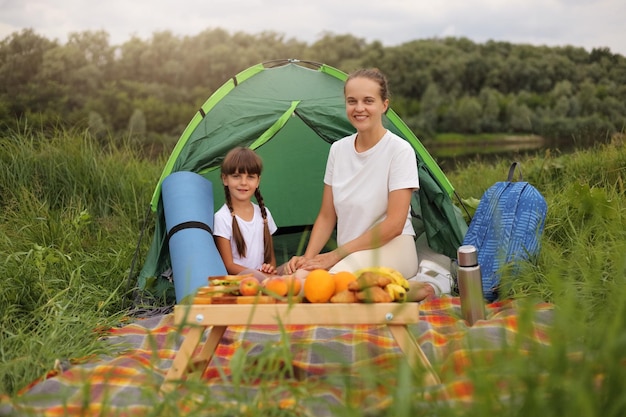 Imagen de una mujer sonriente feliz satisfecha y una niña con pantalones blancos sentadas en una manta cerca de una tienda haciendo un picnic comiendo frutas y sándwiches expresando emociones positivas