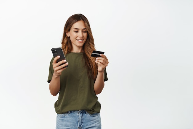 Imagen de una mujer sonriente de mediana edad leyendo el número de la tarjeta de crédito para completar la información en la aplicación de teléfono móvil pagando en línea para hacer compras en el fondo blanco del teléfono inteligente