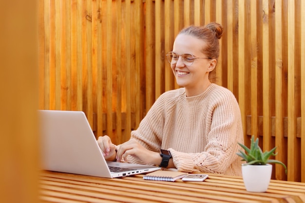 Imagen de una mujer sonriente con peinado de moño con suéter beige y anteojos trabajando en una computadora portátil contra una pared de madera escribiendo en una computadora portátil y mirando la pantalla con una sonrisa con dientes