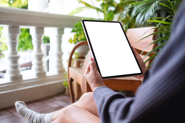 Imagen de una mujer sosteniendo y usando una tableta digital con una pantalla de escritorio blanca en blanco mientras se sienta en el balcón de su casa