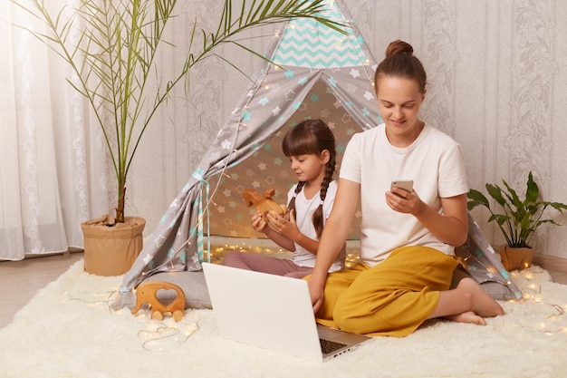 Imagen de una mujer y su hija con camisetas blancas sentadas en un wigwam frente a un niño portátil jugando con una madre de juguete usando un teléfono inteligente y una computadora portátil para trabajar