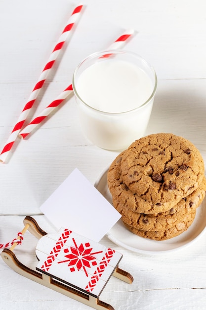 Imagen de navidad con vaso de leche, pajitas a rayas, galletas, nota en blanco, trineo decorativo sobre mesa blanca