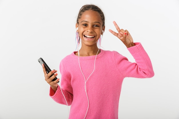 Imagen de una niña afroamericana sonriente sosteniendo un teléfono celular y auriculares mientras gesticula el signo de la paz aislado sobre la pared blanca