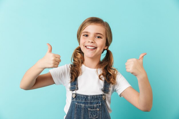 Imagen de una niña bastante joven posando aislada sobre la pared azul mostrando los pulgares para arriba.