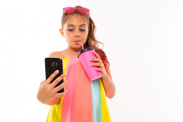 La imagen de una niña caucásica con gafas de sol y teléfono bebe café o jugo y se comunica con sus amigos o familiares
