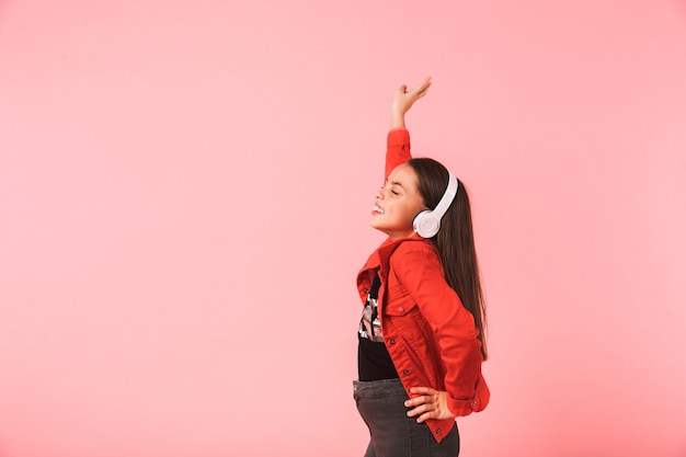 Imagen de la niña feliz en casual con auriculares bailando mientras escucha música, aislado sobre la pared roja