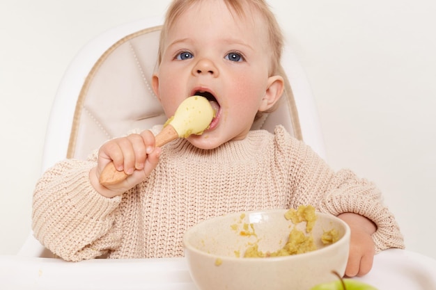 La imagen de una niña graciosa y hambrienta sentada en una silla y comiendo sosteniendo una cuchara en las manos mantiene la boca abierta disfrutando del puré o las gachas usando un jersey beige