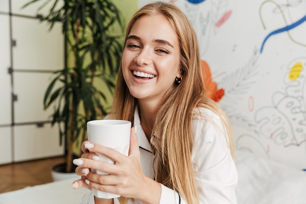 Imagen de una niña linda joven sonriente feliz en pijama en casa en la cama tomando café.