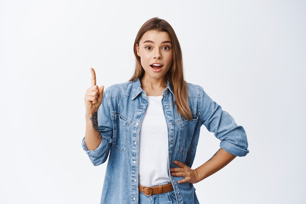 Imagen de una niña milenaria levantando el dedo en un gesto de eureka, lanzando una idea, señalando con el dedo hacia arriba para decir una sugerencia, recomendando un anuncio, pared blanca