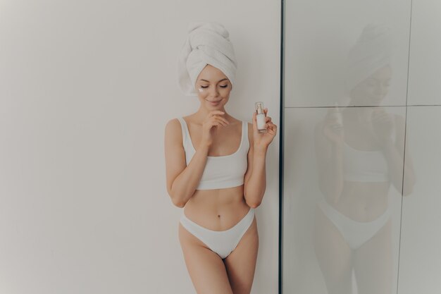 Imagen de niña posando con crema facial hidratante cosmética o suero en la mano aislada sobre pared ligera en el baño, usa una toalla envuelta en la cabeza después de la ducha matutina. Concepto de belleza y cuidado de la piel.