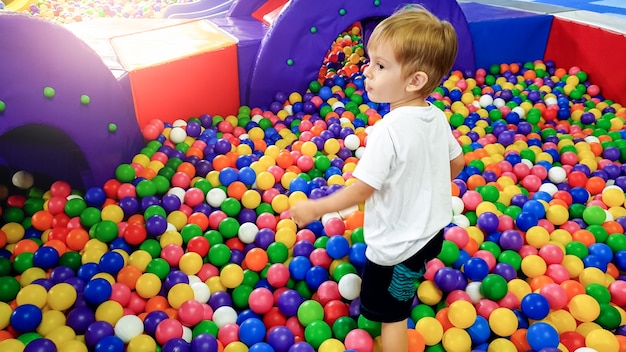 Imagen de un niño de 3 años jugando y divirtiéndose en el patio de recreo con un montón de pequeñas bolas de plastc de colores. Niño disfrutando del parque de atracciones en el centro comercial.