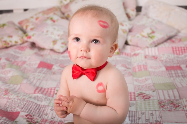 Imagen de niño adorable con besos rojos en la piel, niño feliz, sentado en la cama en el estudio, ángel, vacaciones románticas, día de San Valentín.