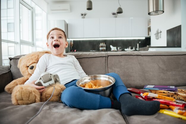 Imagen de niño feliz en el sofá con osito de peluche en casa jugando con la consola mientras come patatas fritas. Sosteniendo el joystick.