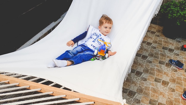 Imagen del niño pequeño adorable sonriente balanceándose y acostado en una hamaca en el patio de la casa