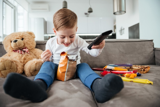 Imagen de niño sentado en el sofá con osito de peluche en casa y viendo la televisión mientras come patatas fritas. Sosteniendo el control remoto.