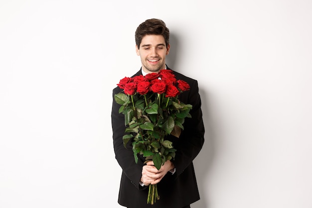 Imagen de novio guapo en traje negro, sosteniendo un ramo de rosas rojas y sonriendo, estando en una cita, de pie sobre fondo blanco.