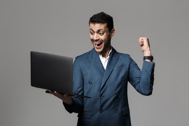 Imagen del optimista hombre árabe de 30 años en traje de negocios sonriendo y sosteniendo un portátil, aislado sobre una pared gris