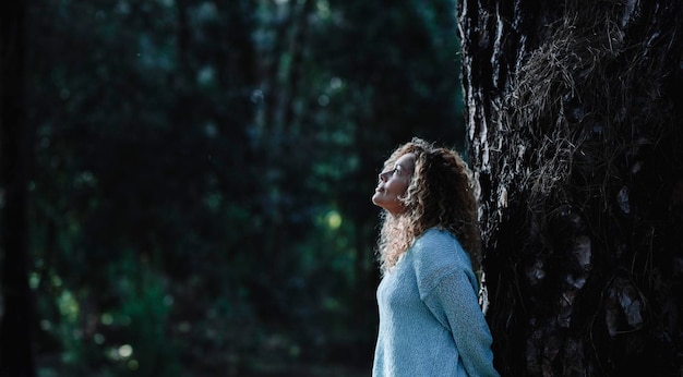 Imagen oscura de una mujer de pie contra el tronco de un árbol en la naturaleza al aire libre actividad de ocio con bosques en el fondo Una mujer disfruta del bosque mirando hacia arriba y relajándose Medio ambiente en el parque Copiar espacio