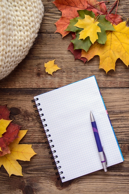 Imagen de otoño con un cuaderno y un bolígrafo, hojas amarillas y bufanda sobre fondo de madera