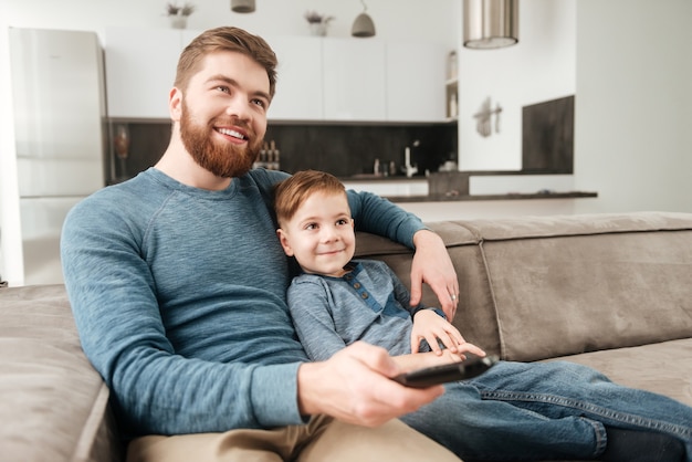 Imagen del padre barbudo feliz que sostiene el control remoto mientras ve la televisión con su pequeño y lindo hijo.