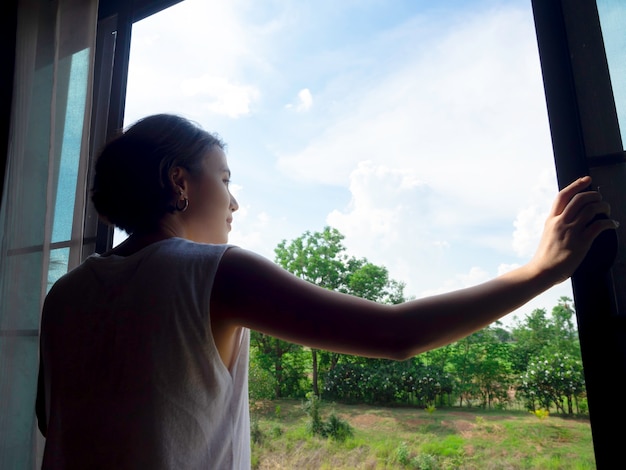 Foto imagen de la parte posterior del pelo corto de la mujer asiática que abre una ventana de cristal grande en el dormitorio en el segundo piso de la casa y que mira el paisaje verde y el cielo azul en un día soleado.