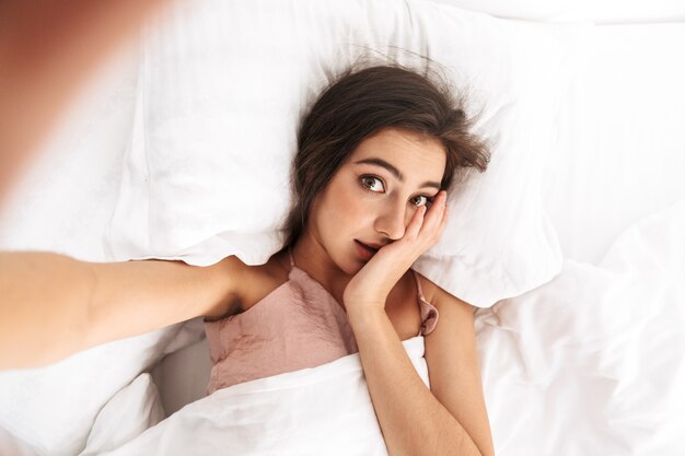 Imagen de la parte superior de una mujer bonita de 20 años con cabello oscuro tomando una foto selfie, mientras está acostada en la cama debajo de la ropa blanca